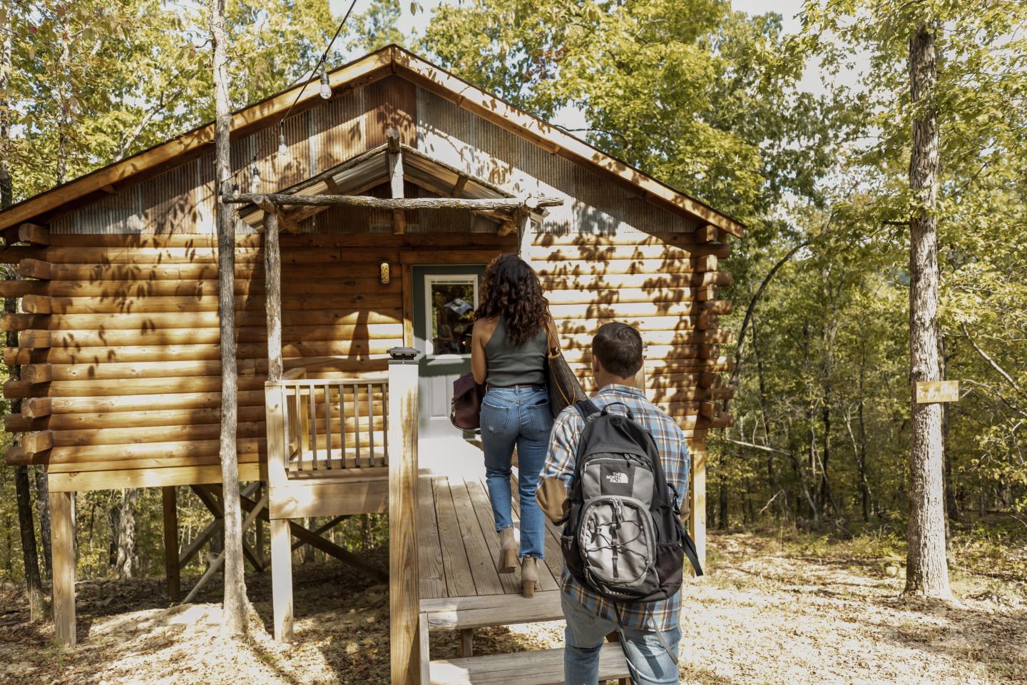 Two people walking up to a wooden cabin in a forest setting.