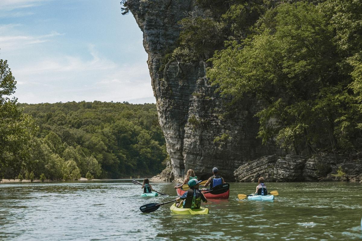 Kayakers paddle on a river surrounded by lush trees and rocky cliffs.