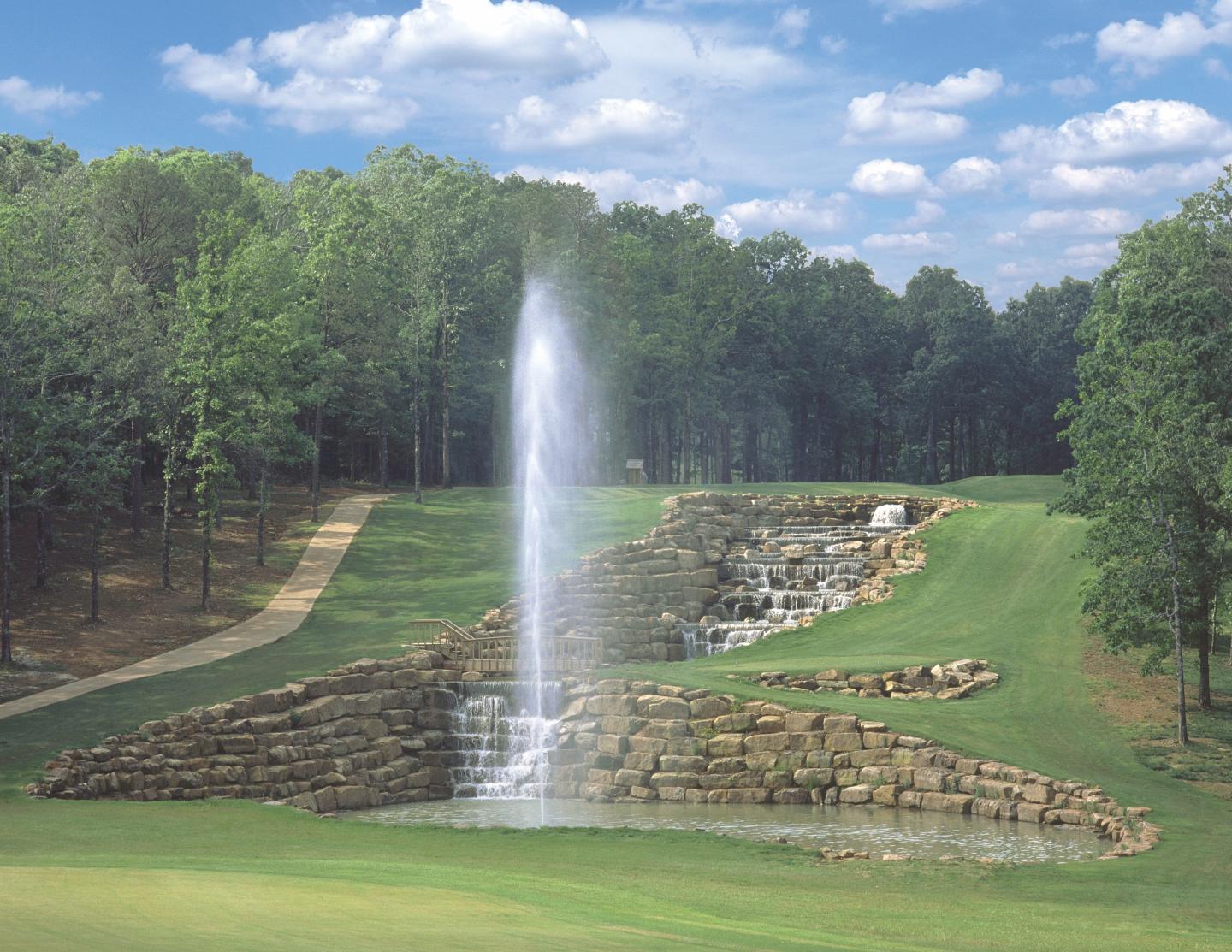 Water fountain amidst a lush green park with trees and blue sky.