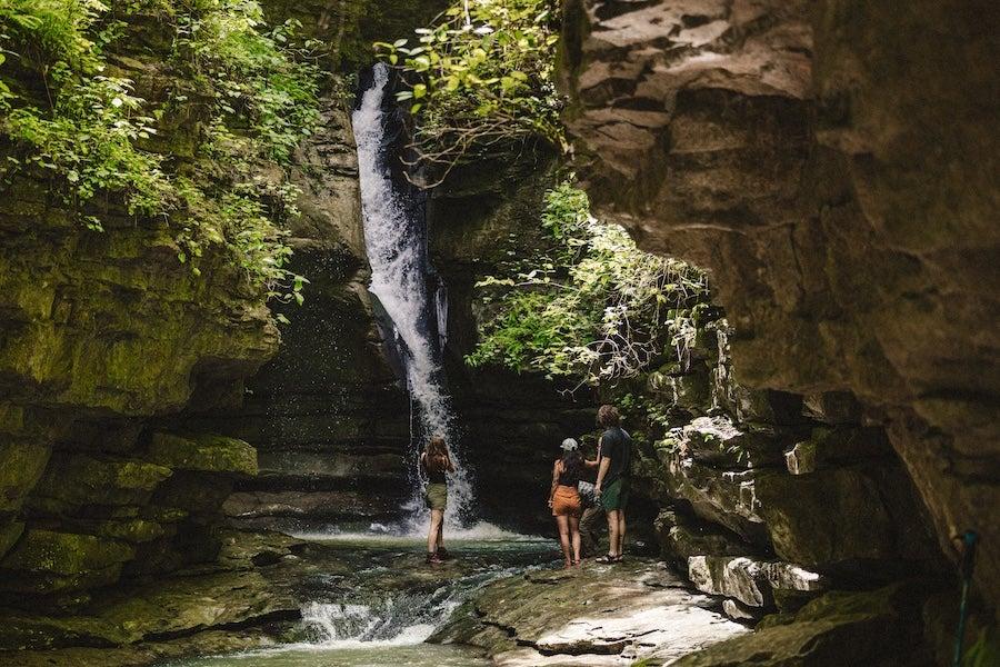 Three people stand by a waterfall in a lush, rocky gorge.