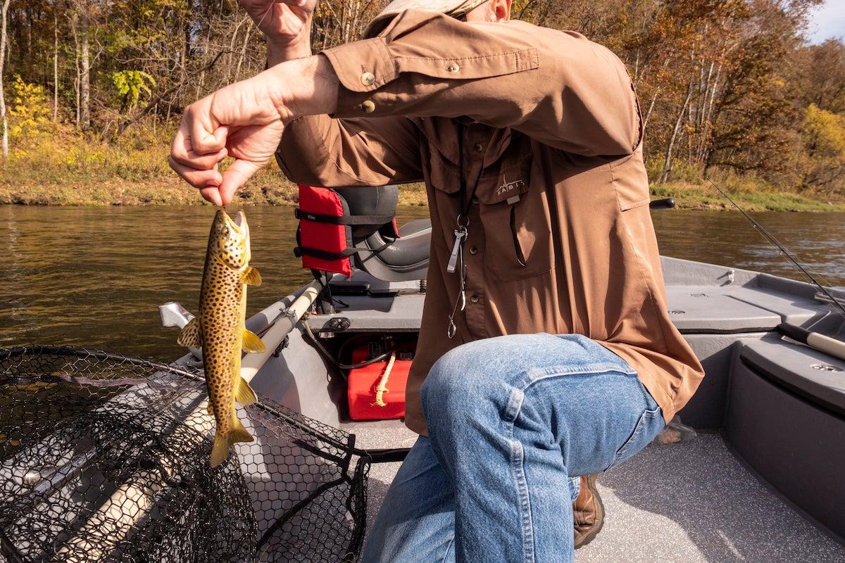 Man kneeling on boat holding a small fish by the water.