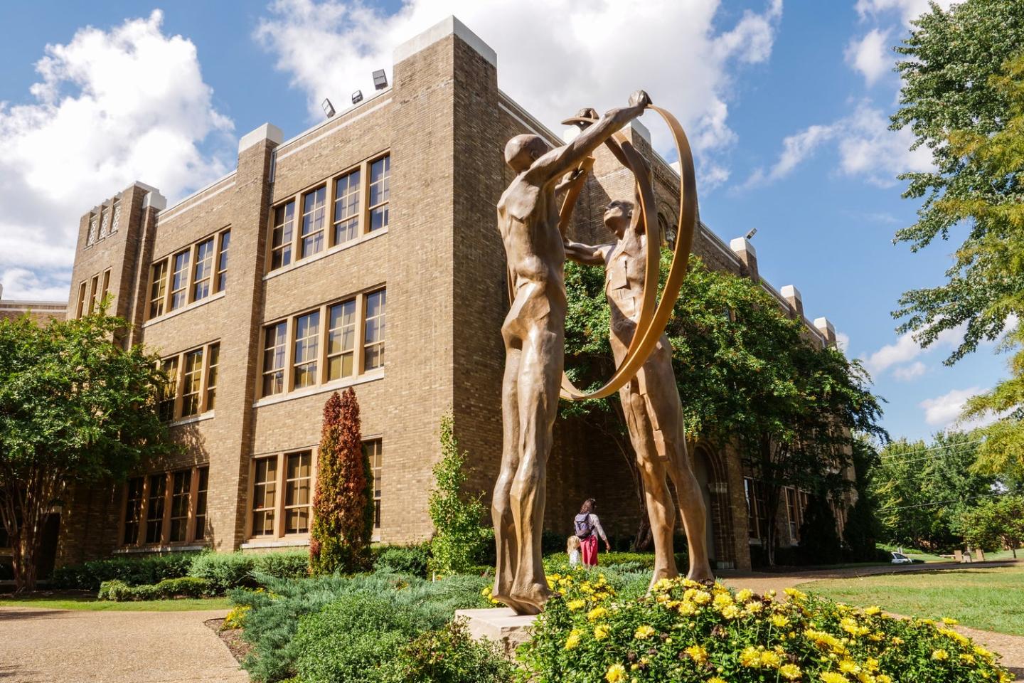 Bronze sculpture of two figures with an arch, building and garden in the background.