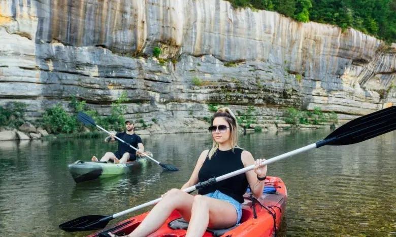 Two people kayaking on a calm river near a rocky cliff, one in red, one in green.