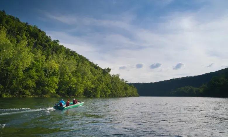 Kayakers on a calm river, surrounded by lush green hills under a blue sky.