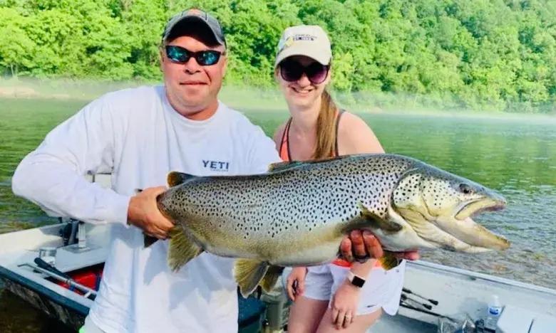 Two people on a boat holding a large fish, with trees in the background.