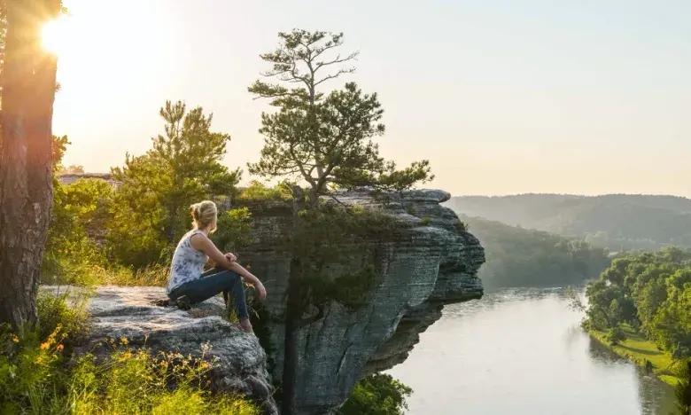 Person sitting on a cliff edge, overlooking a river at sunset.