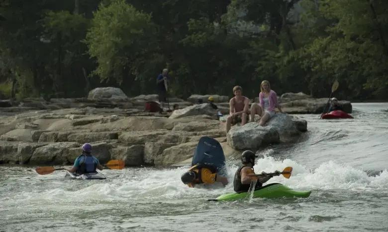 Kayakers navigating river rapids as people watch from rocks.