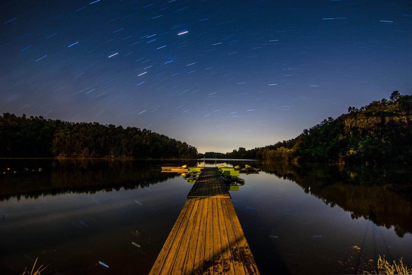 Wooden pier on calm lake under starry night sky.