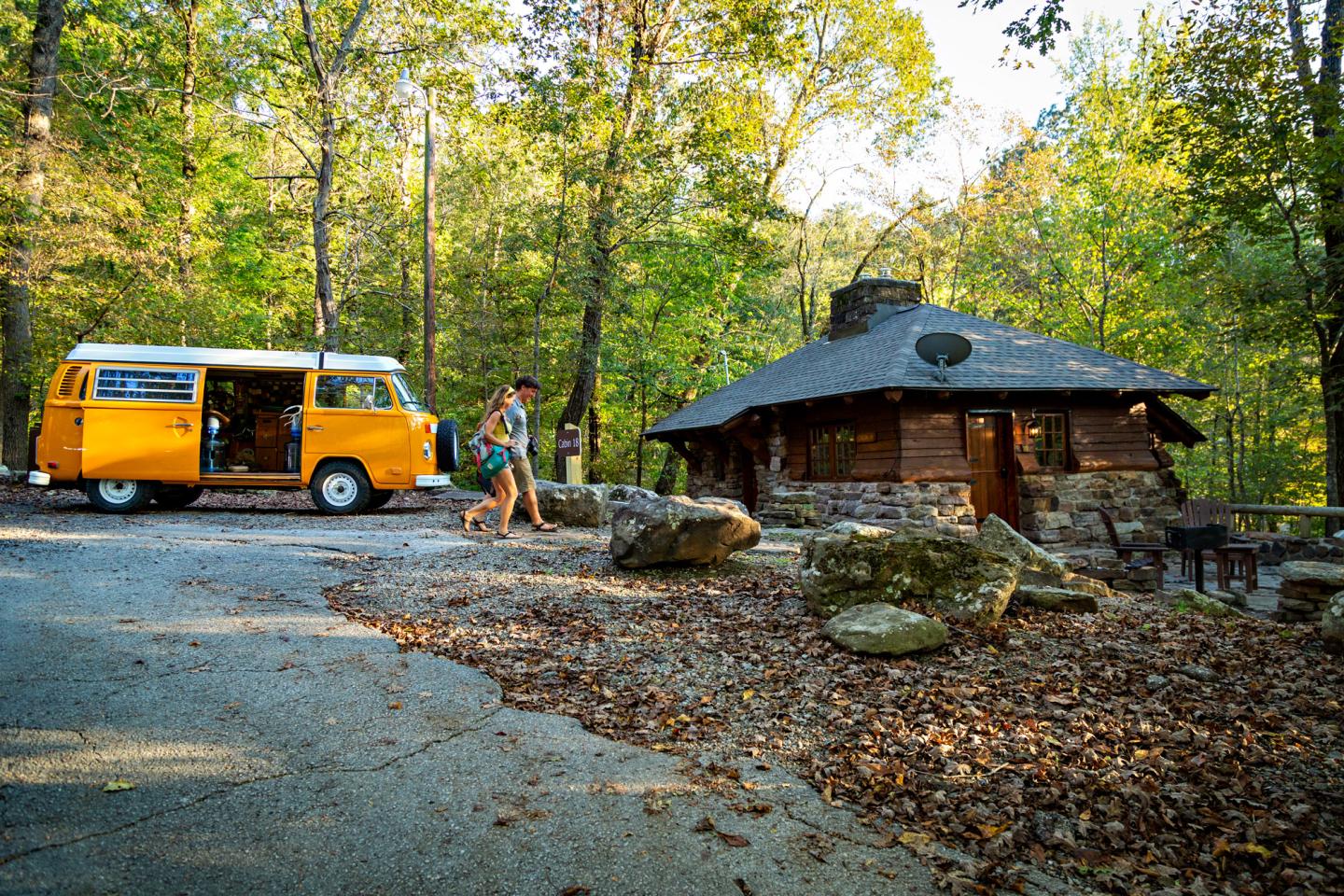 Yellow camper van and cabin in wooded area with two people walking..