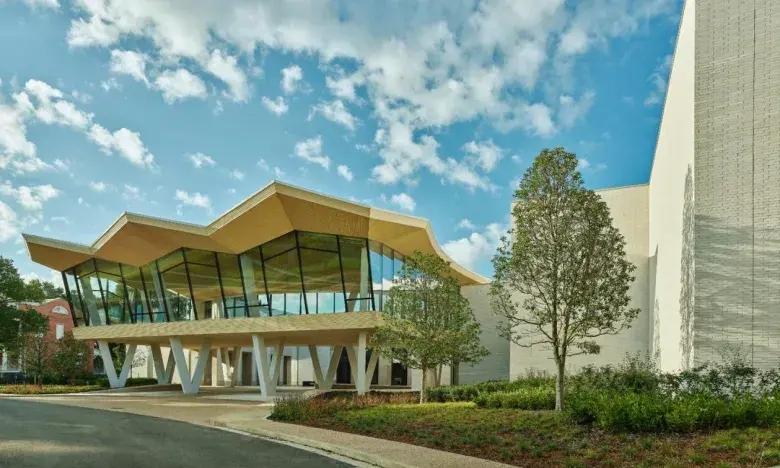 Modern building with zigzag roof and large windows, surrounded by trees and cloudy sky.