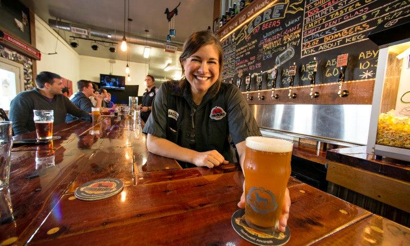 Bartender smiling, holding a glass of beer at a lively bar counter.