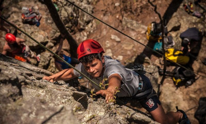 Boy rock climbing, wearing a red helmet, reaching for a hold.