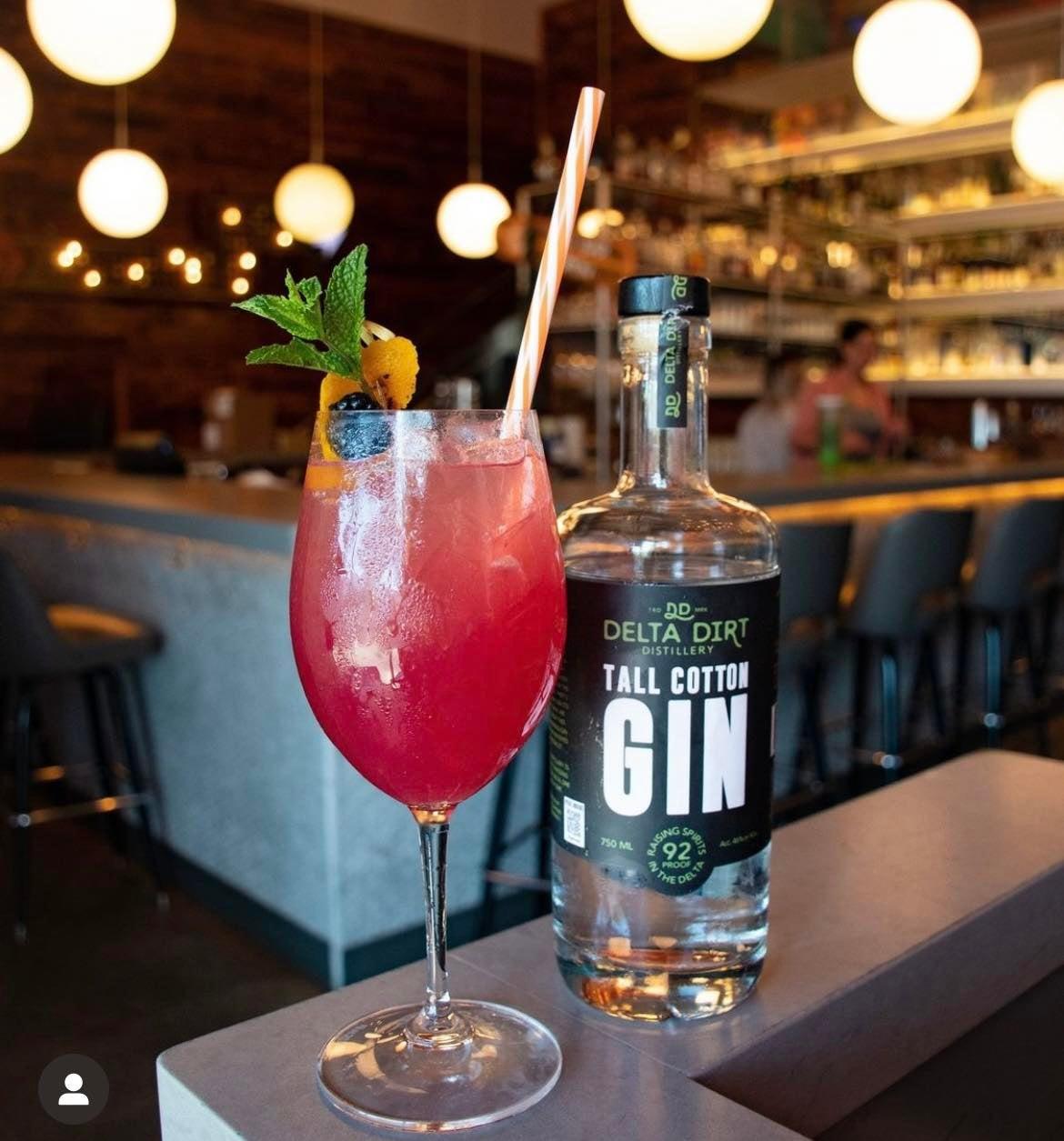 Red cocktail with fruit garnish and gin bottle on bar counter, warm lighting.