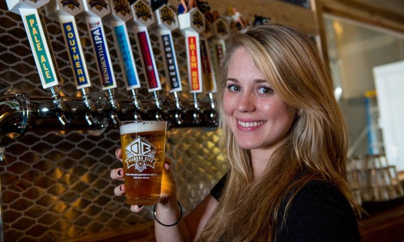 Woman holding a beer glass, smiling in front of a bar with multiple tap handles.