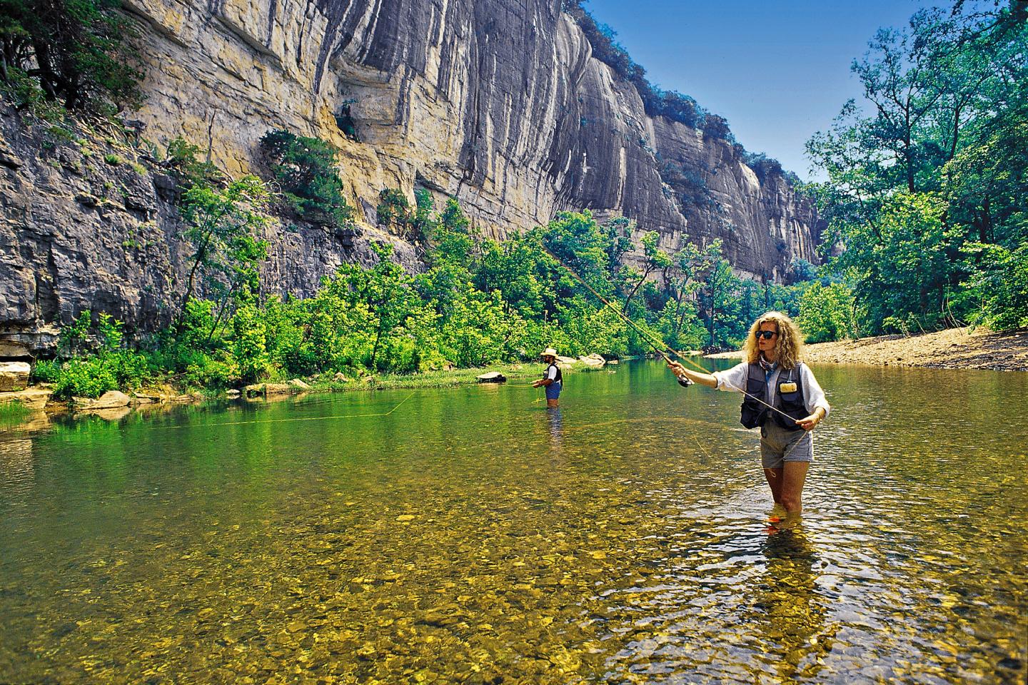 Two people fly fishing in a clear, shallow river with rocky cliffs and lush greenery.
