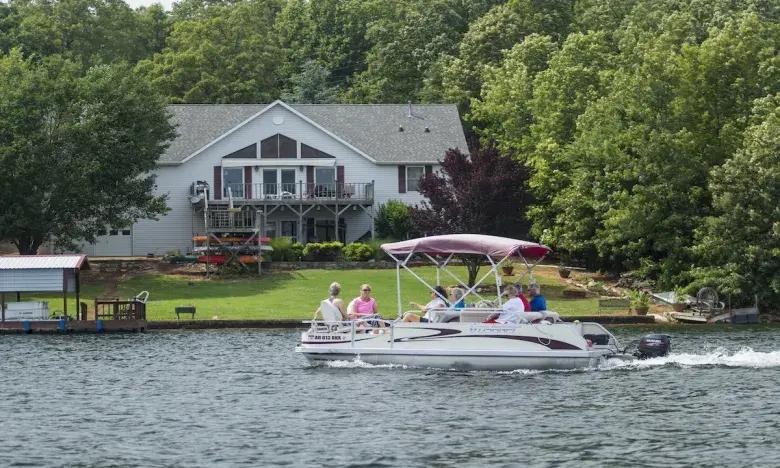 Pontoon boat rides on a lake near a house surrounded by trees.
