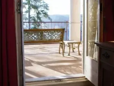 Open door leading to a sunlit balcony with a bench and table. Forested view in the distance.
