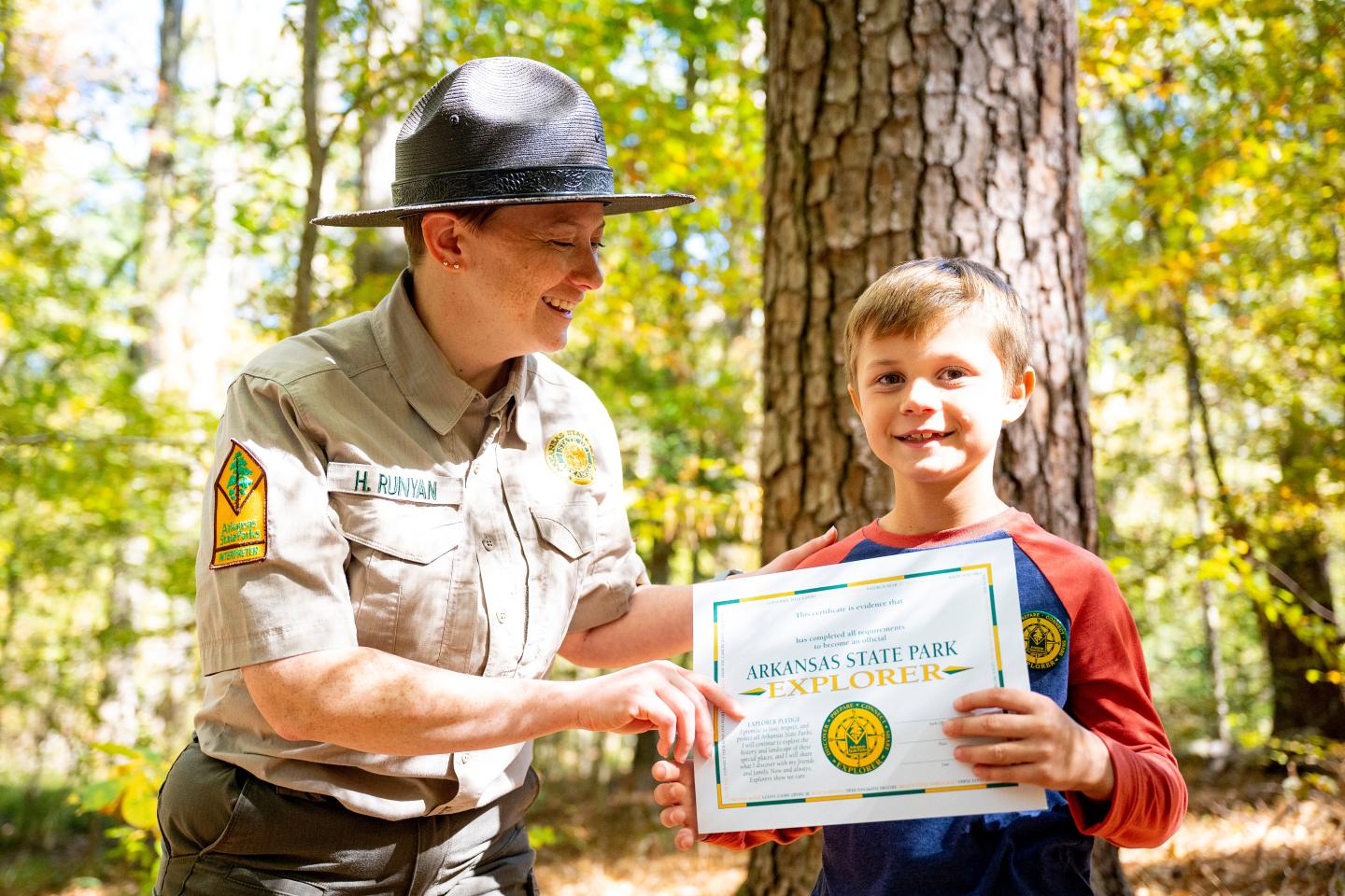 Park ranger and boy smiling with a certificate in a sunny forest.