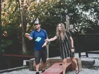Man and woman playing cornhole outdoors, trees in the background.