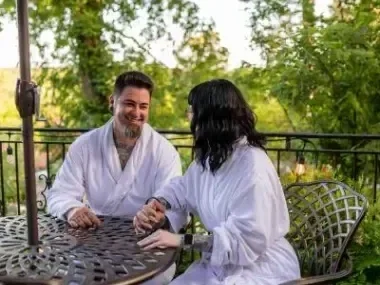 Couple in white robes smiling at each other on a patio surrounded by greenery.