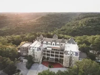 Historic castle-like building surrounded by lush green forest at sunset.