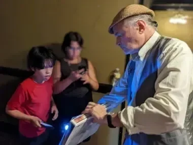 Older man with cap interacting with two kids using gadgets, in dim lighting.