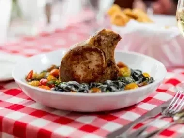 Grilled pork chop on pasta with colorful vegetables, checkered tablecloth.