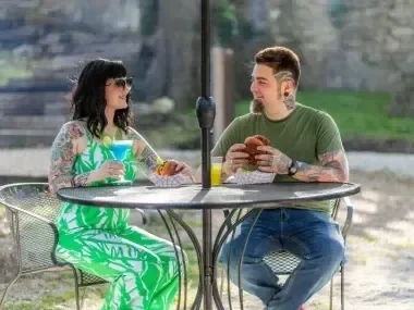 Two people with tattoos seated at an outdoor café table, enjoying drinks and food.