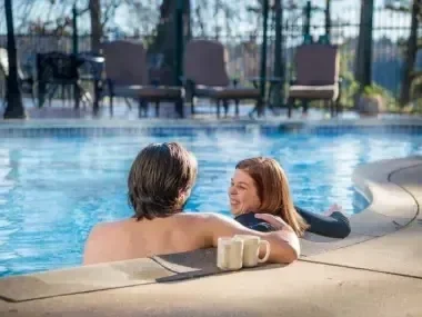 Couple smiling in a pool, drink on edge, chairs in background.