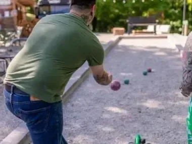 Man playing bocce ball on a gravel court, surrounded by greenery.