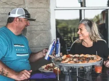 A man and woman smiling over a large pizza on a patio.