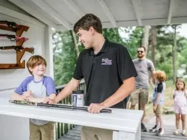 Young boy and man at a shooting range counter, with guns on display, outdoor setting.