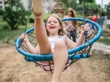 A child joyfully swinging on a round blue net swing in a park.