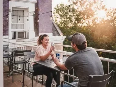 Two people laughing on a sunny balcony, holding hands over a table.