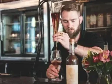 Bartender pouring drink into glass, with flowers and a bottle on the counter.