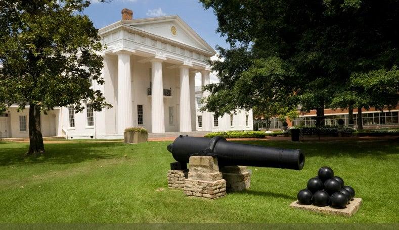 White-columned building with a cannon in front on a grassy lawn.