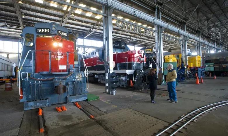 Trains displayed in a large indoor railway depot with two people walking nearby.