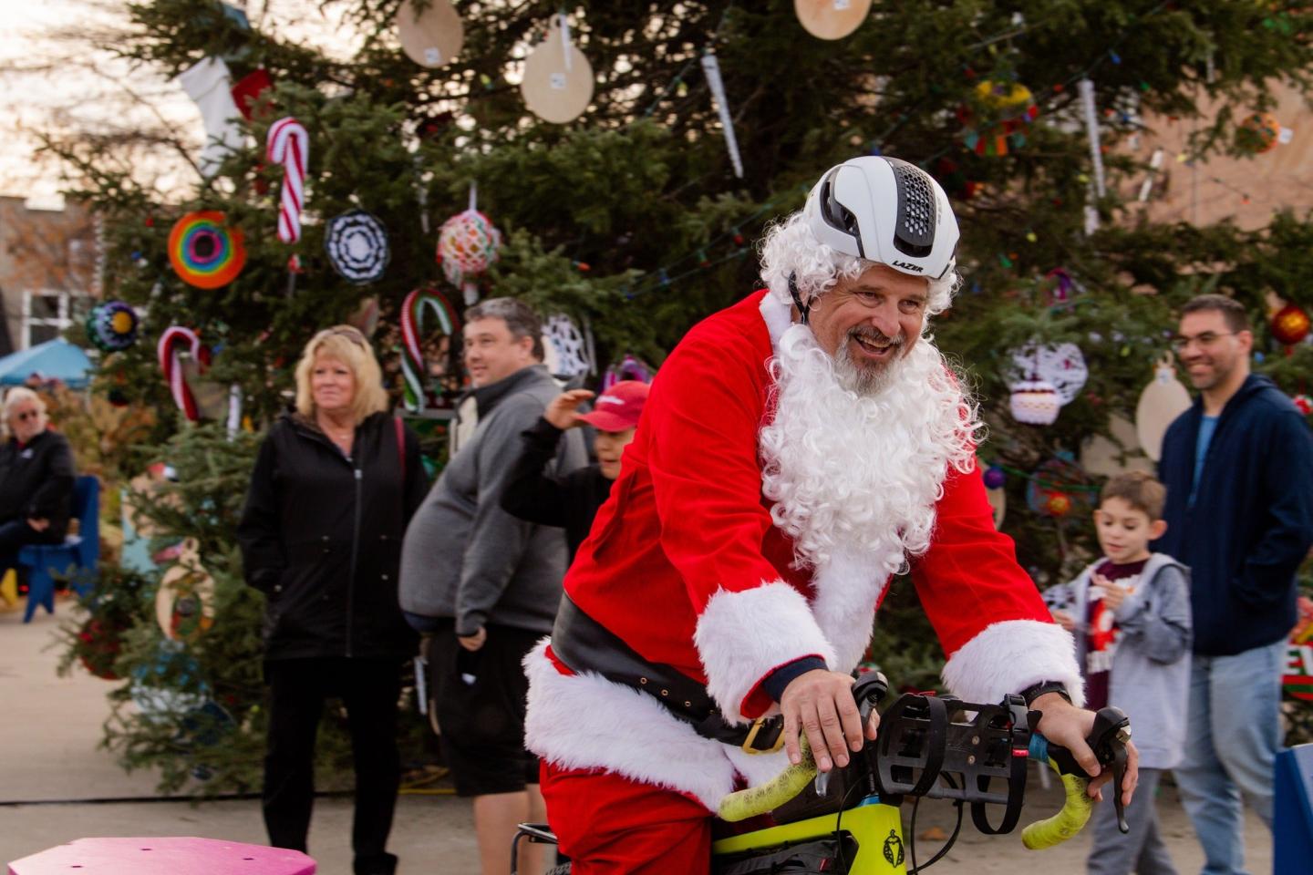 Santa riding a bike in front of a decorated Christmas tree, people watching.