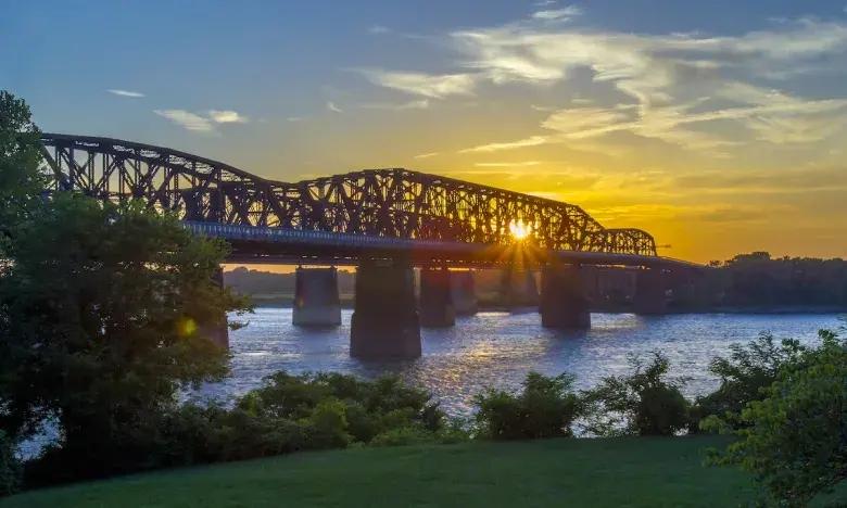 Sunset over river with a bridge, framed by trees on grassy banks.