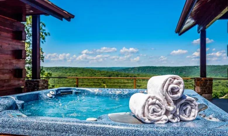 Hot tub on deck with rolled towels overlooking a scenic forest landscape under a clear blue sky.
