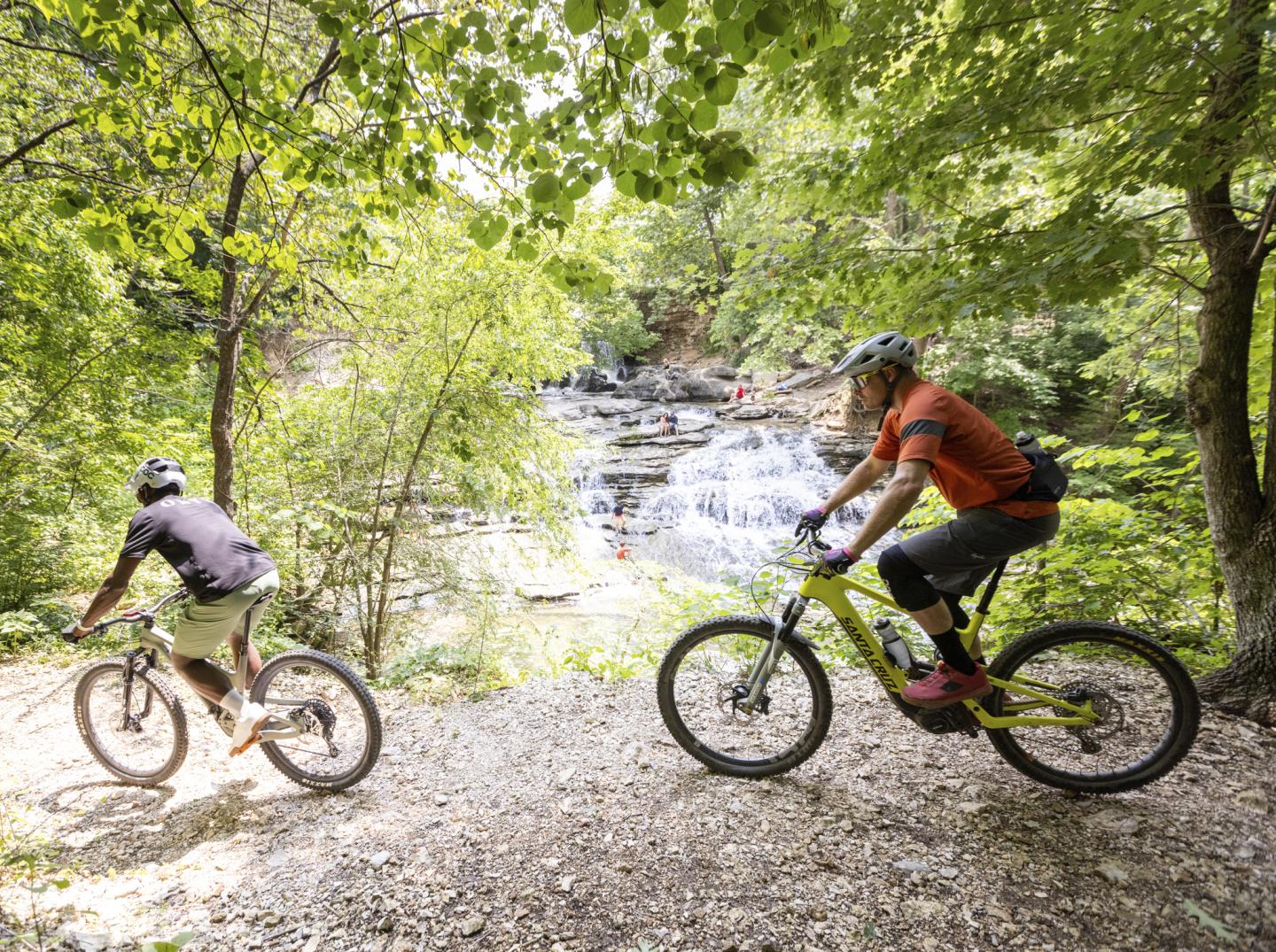 Cyclists on a forest trail near a waterfall.