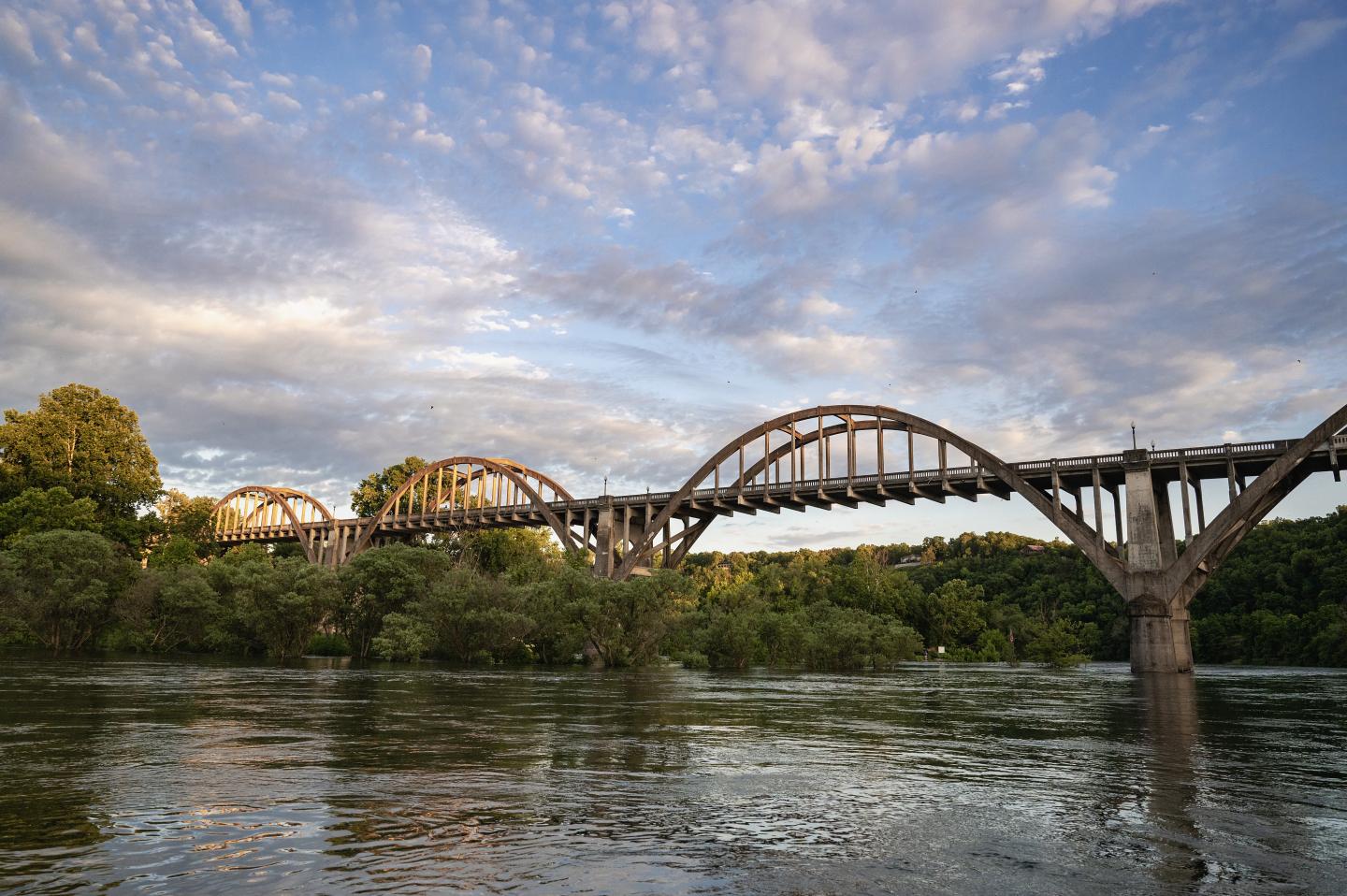 Arched bridge over a river with trees and a cloudy sky.