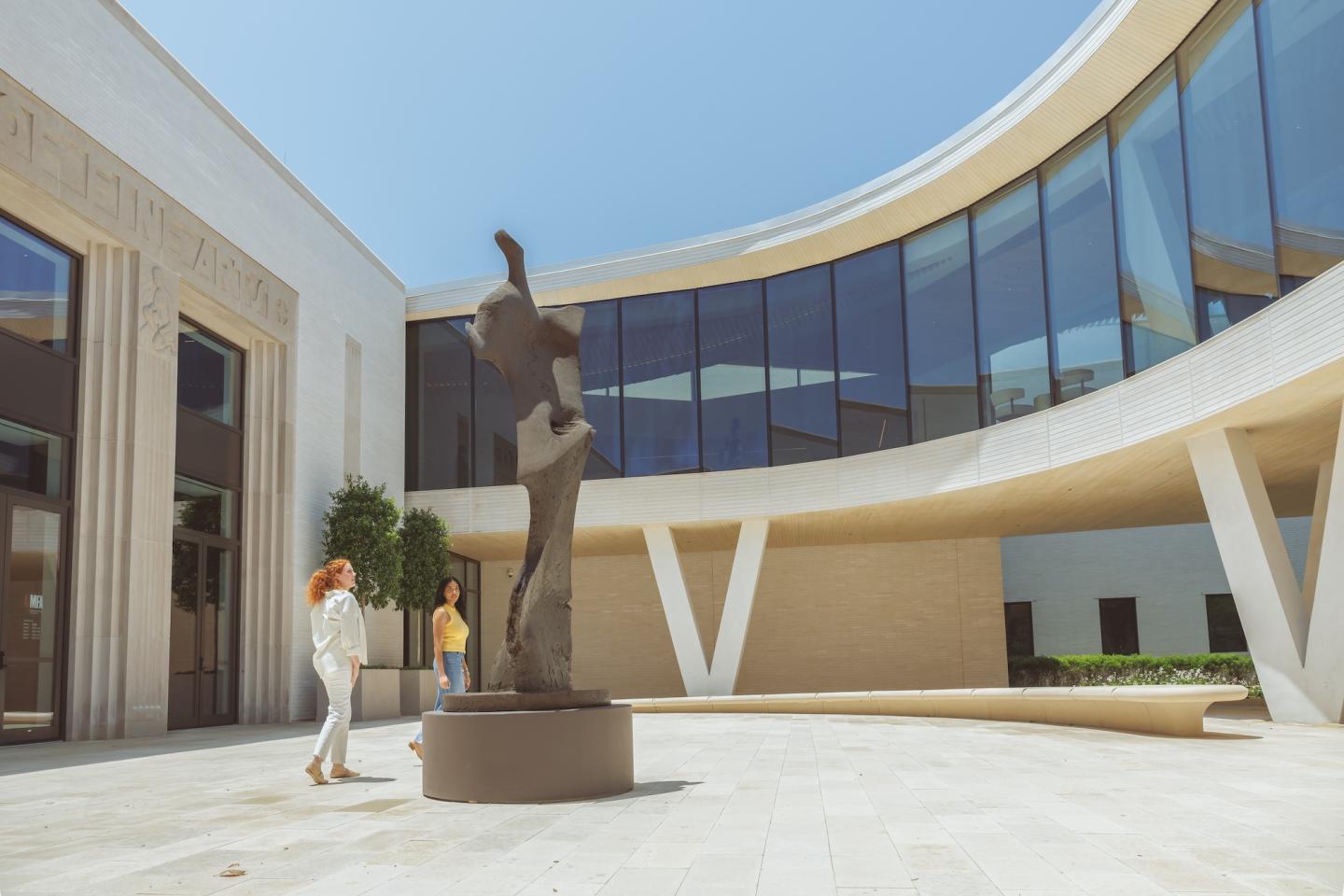 Curved building courtyard with abstract sculpture and two people in sunlight.