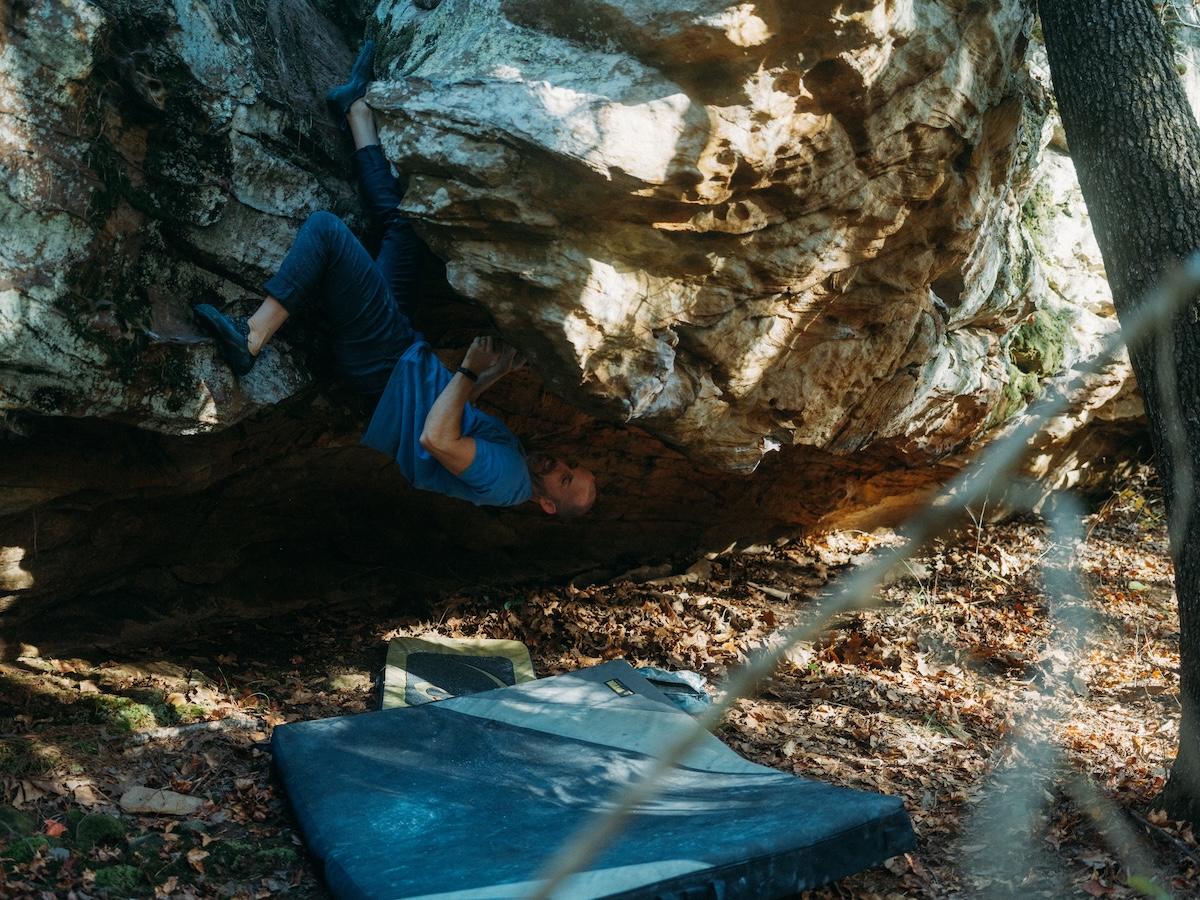Climber in a blue shirt bouldering under a large rock, with crash pads below.