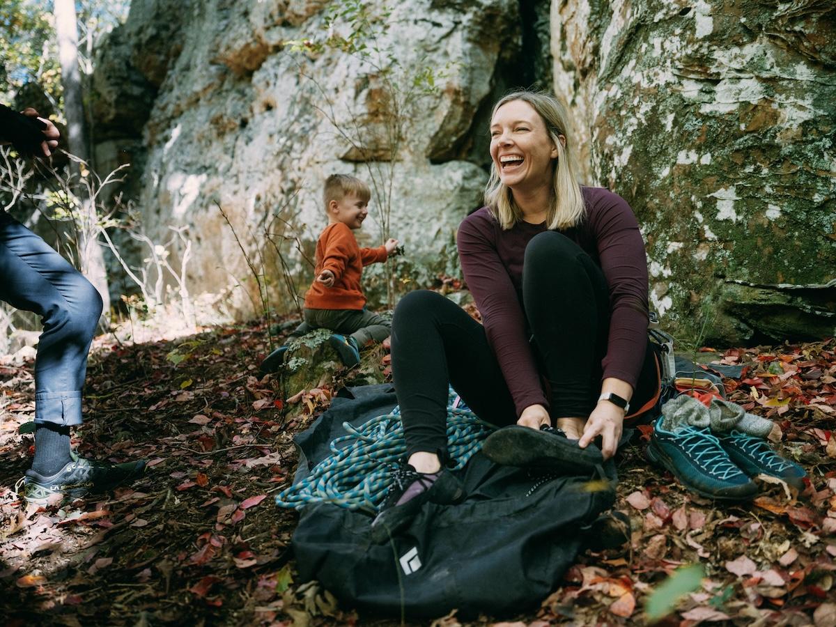 Woman laughing while sitting on a blanket outdoors, with child in background.