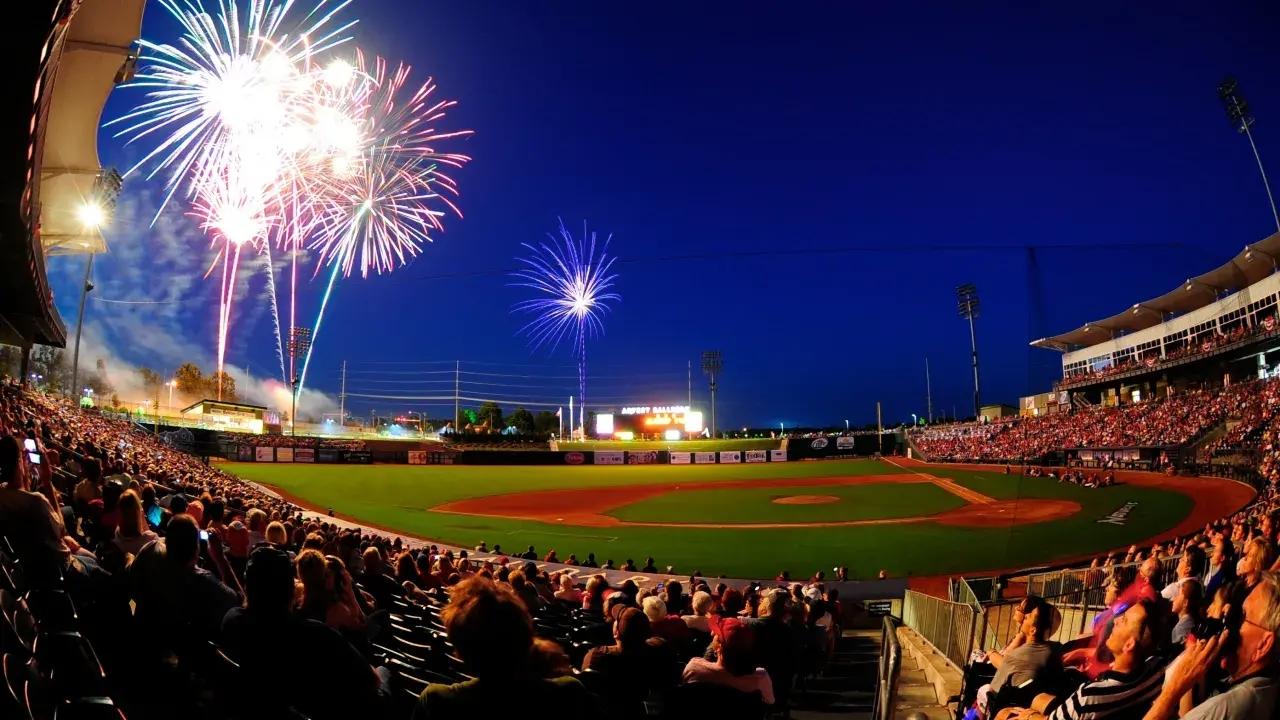 Fireworks light up the sky over a crowded baseball stadium at night.