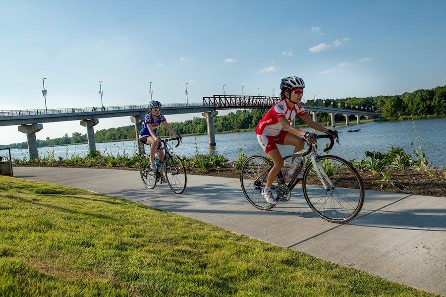 Cyclists ride along a riverside path under a clear blue sky.
