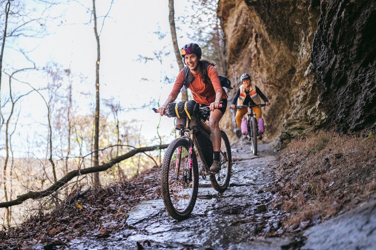 Cyclists ride along a rocky, wooded trail wearing safety gear.