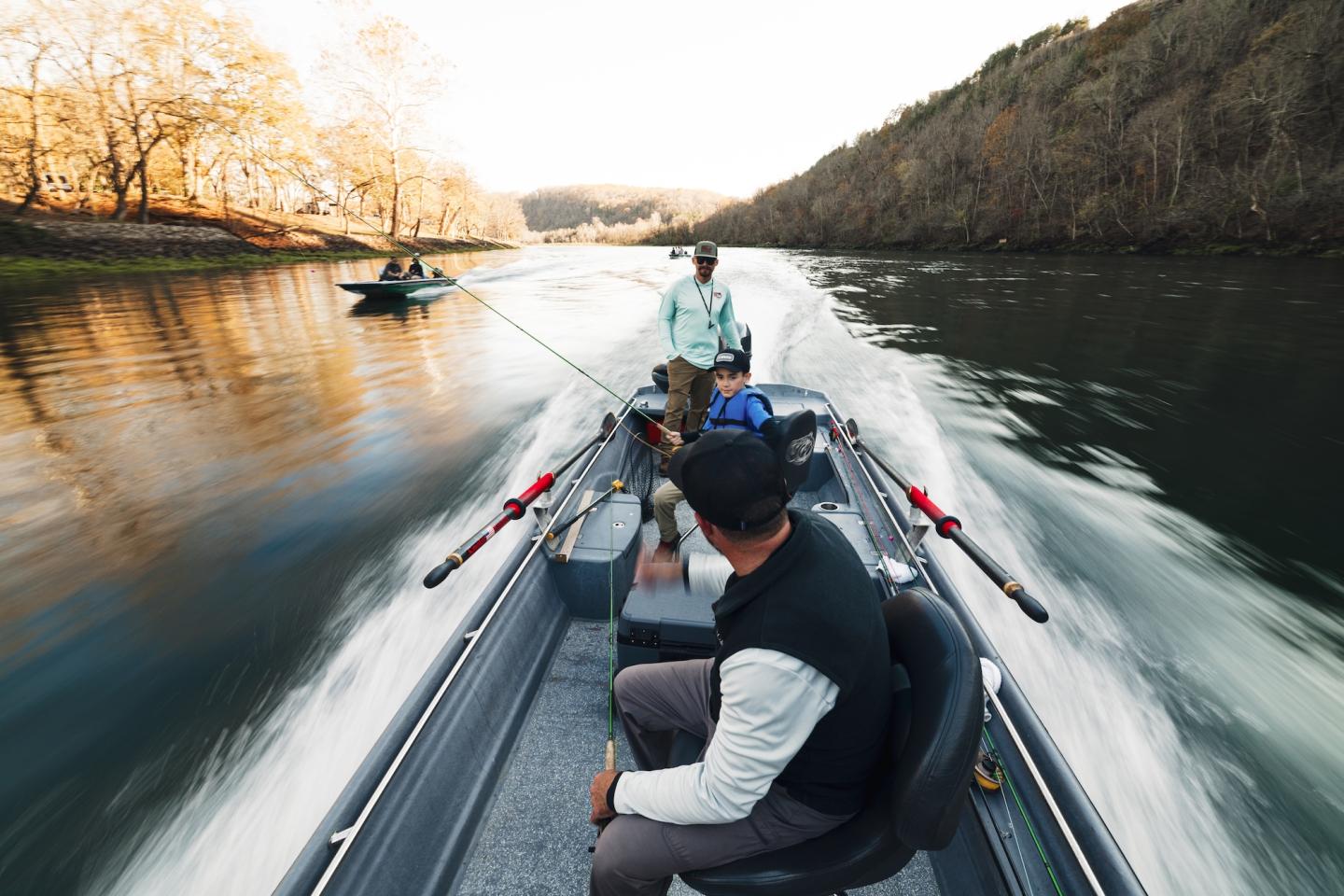 Two people fishing on a river in a motorboat, surrounded by trees.