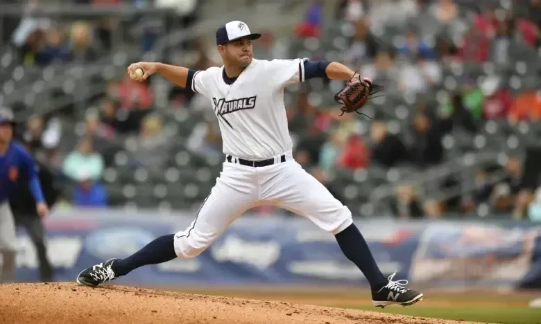 Baseball player pitching in a stadium, wearing a white uniform and cap.